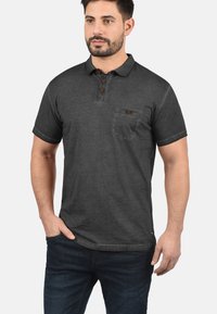 Mørkegrå polo-shirt med krave, tre brune knapper og en lomme på venstre bryst. Korte ærmer og en afslappet pasform.