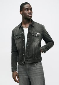 Čierna denimová bunda s klasickým strihom, vybavená dvoma náprsními vreckami, striebornými gombíkmi a mierne vyblednutým vzhľadom. Nosená cez bielu košeľu.