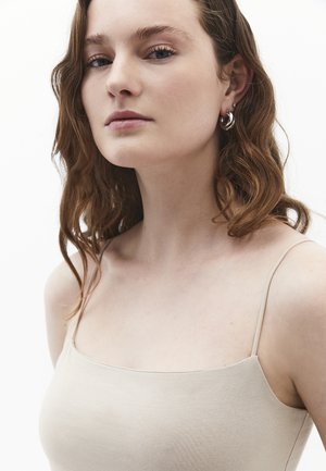 Débardeur beige à fines bretelles, avec une texture lisse et un col carré. Le mannequin porte des boucles d'oreilles simples en argent.
