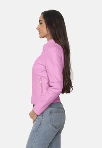 Chaqueta de imitación de cuero rosa con un diseño ajustado, cuello alto y bolsillos laterales con cremallera. Manos largas y textura suave visibles.