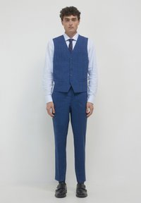 Giovane uomo in piedi, con un gilet e pantaloni a quadri blu, camicia bianca, cravatta scura e scarpe monk nere a doppia fibbia, su sfondo semplice.