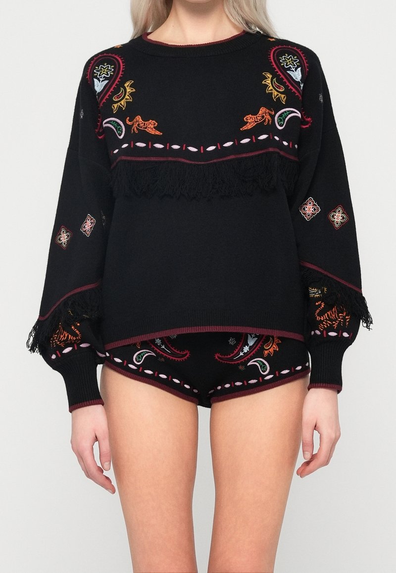 Femme portant un pull à manches longues noir et un short assorti avec des motifs brodés colorés et des détails en franges.