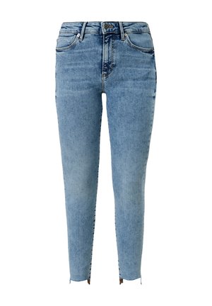 Vrouw draagt een witte top met pofmouwen, lichtblauwe high-waisted skinny jeans en witte sneakers, staand met handen in de zij.