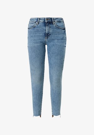 Kvinde iført hvid bluse med puffede ærmer, lyseblå højtaljede skinny jeans og hvide sneakers, stående med hænderne på hoften.