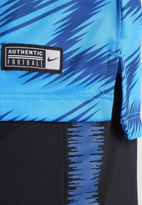 Camiseta deportiva azul con patrones en zigzag en distintos tonos, acentos negros y una etiqueta que dice "FÚTBOL AUTÉNTICO" de Nike.