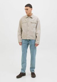 Jachetă din denim bej deschis cu două buzunare pe piept, nasturi metalici argintii și o croială lejeră, asortată cu blugi albăstrui și pantofi maro închis.