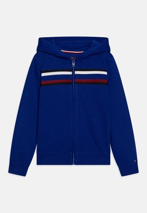 Modrá mikina na zip s kapucí, s vodorovnými pruhy v červené, bílé a černé barvě přes hrudník. Měkký materiál s žebrovanými manžetami.