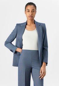 Maßgeschneiderte blaue Blazer mit Notch-Revers, Ein-Knopf-Verschluss und Dreiknopf-Manschetten, kombiniert mit einem hellen gerippten Tanktop und passenden Hosen.