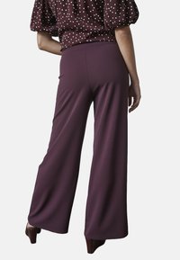Vida byxor i en djup lila tyg, med en slät yta och elastiskt midjeband, kombineras med en bourgogne-färgad blus med prickar.