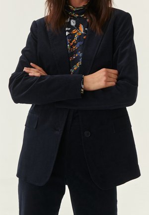 Blazer en velours côtelé navy avec deux poches avant, fermeture à un bouton et revers structurés. Porté sur un col roulé à motifs.
