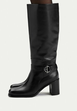 Schwarze kniehohe Lederstiefel mit Blockabsätzen und einem silbernen runden Schnallendetail am Knöchelriemen, getragen mit schwarzen Strümpfen.