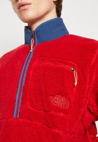 Pulóver de forro polar rojo con cuello azul, media cremallera y bolsillo en el pecho con cremallera. Cuenta con un logo en relieve y tejido texturizado. Acento en cordón amarillo.