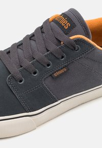 Gray canvas low-top sneaker with orange accents. Features metal eyelets, flat gray laces, and a white rubber sole with a textured finish.