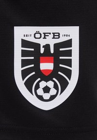 Ασπρόμαυρο έμβλημα σε σχήμα ασπίδας με στυλιζαρισμένο αετό, κόκκινο και άσπρο ριγωτό οικόσημο, μπάλα ποδοσφαίρου και το κείμενο "SEIT ÖFB 1904".