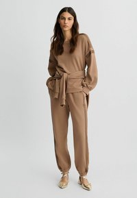 Braunes Athleisure-Set mit einem Langarmoberteil mit gebundenem Bund und passenden tapered Pants. Glatte Textur mit seitlichen Akzenten. Goldene flache Schuhe.