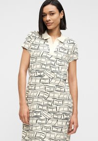 Γυναίκα με ίσια σκουρόχρωμα μαλλιά, ντυμένη με κοντομάνικο κρεμ φόρεμα με μαύρο αφηρημένο γραμμικό σχέδιο και μπεζ γιακά, στέκεται μπροστά σε απλό φόντο.