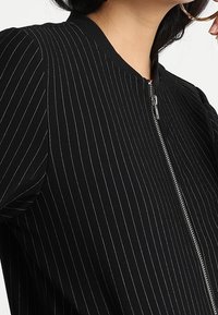 Czarna kurtka z pionowymi białymi paskami, okrągłym dekoltem i zamkiem błyskawicznym z przodu. Materiał wydaje się gładki z delikatnym połyskiem.