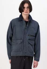 Denim jacket in dark blue with a structured collar, three pockets, and button closures. Features a zippered chest pocket and textured fabric.
