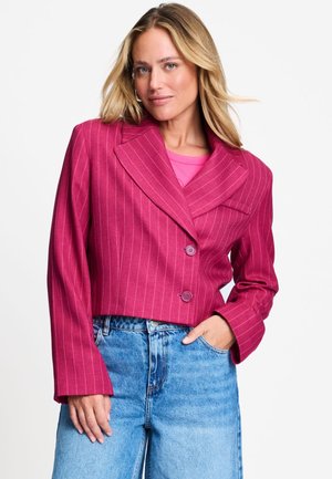 Vrouw met blond haar die een roze krijtstreep cropped blazer draagt over een roze top en blauwe high waist jeans, hand in de zak, neutrale achtergrond.