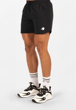 Male legs wearing black athletic shorts with a white gorilla logo, white socks with black stripes, and white and black running shoes.