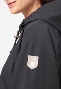 Chaqueta negra impermeable con un acabado texturizado, que cuenta con una capucha, cordones y un parche de logo beige en la manga.
