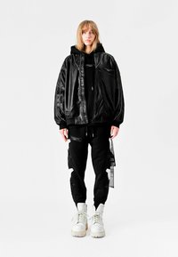 Chaqueta bomber de cuero sintético negro con puños acanalados, con una sudadera negra debajo. Combinada con pantalones jogger negros que presentan parches y detalles desgastados. Botas blancas.