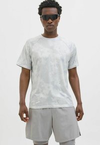 Harmaa urheilupaita, jossa on maastokuvio, lyhyet hihat ja pyöreä pääntie, yhdistettynä vaaleanharmaoisiin shortseihin. Tyylikkään muotoiset aurinkolasit.