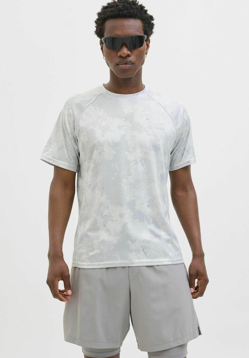 Harmaa urheilupaita, jossa on maastokuvio, lyhyet hihat ja pyöreä pääntie, yhdistettynä vaaleanharmaoisiin shortseihin. Tyylikkään muotoiset aurinkolasit.