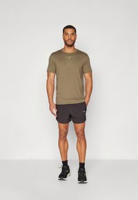 T-shirt desportivo verde-oliva com mangas curtas, combinado com calções pretos. O modelo usa sapatilhas pretas com solas brancas.