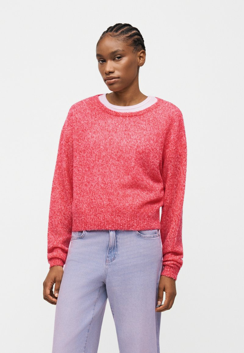 Νεαρή γυναίκα με πλεγμένα μαλλιά, που φοράει ένα ροζ πλεκτό πουλόβερ πάνω από λευκό πουκάμισο και ανοιχτόχρωμα μπλε τζιν, στέκεται μπροστά σε απλό φόντο.