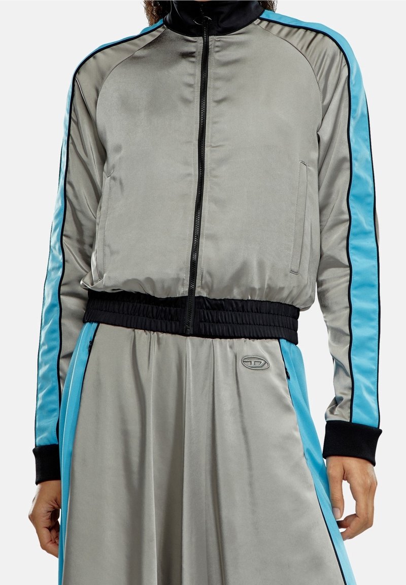 Grå satin zip-jacka med blå sidostråkar och svarta detaljer; har en ribbad svart midja och logotypdetalj på vänster sida.