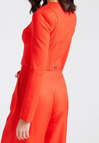 Rode jumpsuit met lange mouwen, getailleerde pasvorm, subtiele studs langs de halslijn en een gestrikte taille-detail. Soepele stof met een lichte glans.