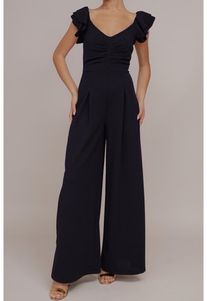 Kvinde iført en sort jumpsuit med flæset korte ærmer, en rynket V-hals top, bukser med vide ben og gyldne sandaler med åben tå.