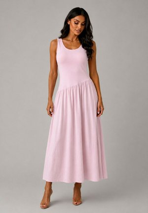 Femme portant une robe longue rose clair sans manches avec taille froncée, debout devant un fond gris uni, posant avec les mains détendues.