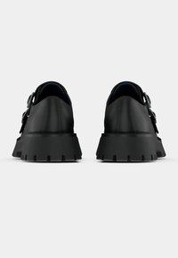 Μαύρα δερμάτινα παπούτσια με χοντρό τακούνι, με υφή στην επιφάνεια και φαρδύ λουρί με αγκράφα. Σχεδιασμένα με στρογγυλεμένη μύτη και ανοίγματα διακόσμησης.