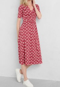 Rotes Midi-Kleid mit weißem Blumenmuster, kurzen Ärmeln, rundem Ausschnitt, gerafftem Taillenbereich und kombiniert mit weißen Sneakers. Glatte Stofftextur.