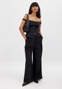 Schwarzes Off-Shoulder-Peplum-Top mit Knopfverschluss und gerüschten Trägern, kombiniert mit hochgeschnittenen, weitem Hosen und spitzen schwarzen Stöckelschuhen.