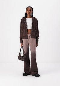Felpa marrone con zip, crop top bianco, jeans a zampa d'elefante marrone chiaro con finitura sfumata, scarpe platform nere e una piccola borsa scura.