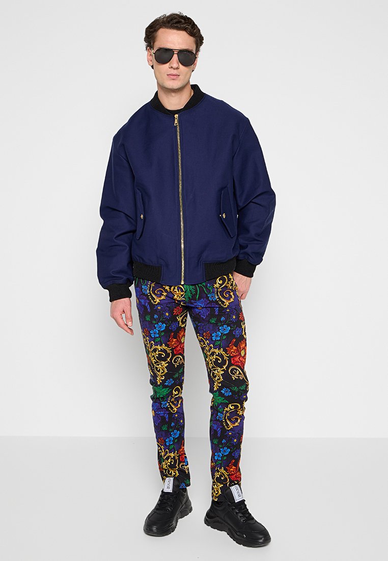 Marineblauer Bomberjacke mit goldenem Reißverschluss, kombiniert mit blütenmusternden Hosen in lebhaften Farben. Schwarze Sneakers runden das Outfit ab.