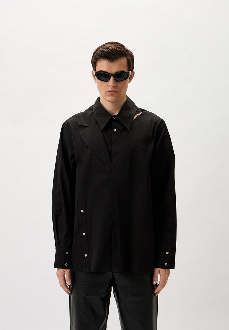 Schwarzes Langarmshirt mit weitem Kragen, Knöpfen vorne und einem Ausschnittdetail an der rechten Schulter. Glatte Stoffqualität mit lockerer Passform.