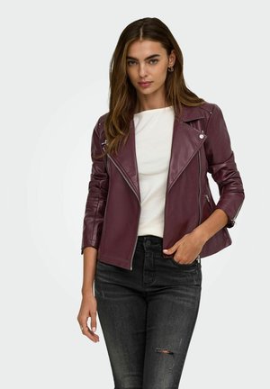 Femme portant une veste en cuir bordeaux par-dessus un haut blanc et un jean noir déchiré, debout avec une main dans la poche devant un fond clair uni.