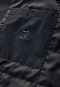 Čierna látka so všitou štítkom s označením "NEXT", veľkosť "M" a "Made in China" na prešívanom textilnom pozadí.