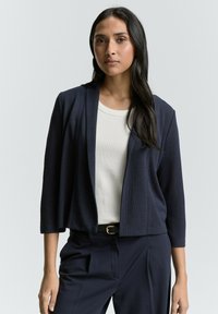 Blazer cropped di colore blu navy con maniche a tre quarti, tessuto texturizzato, design aperto davanti, abbinato a una maglietta bianca e pantaloni blu navy.