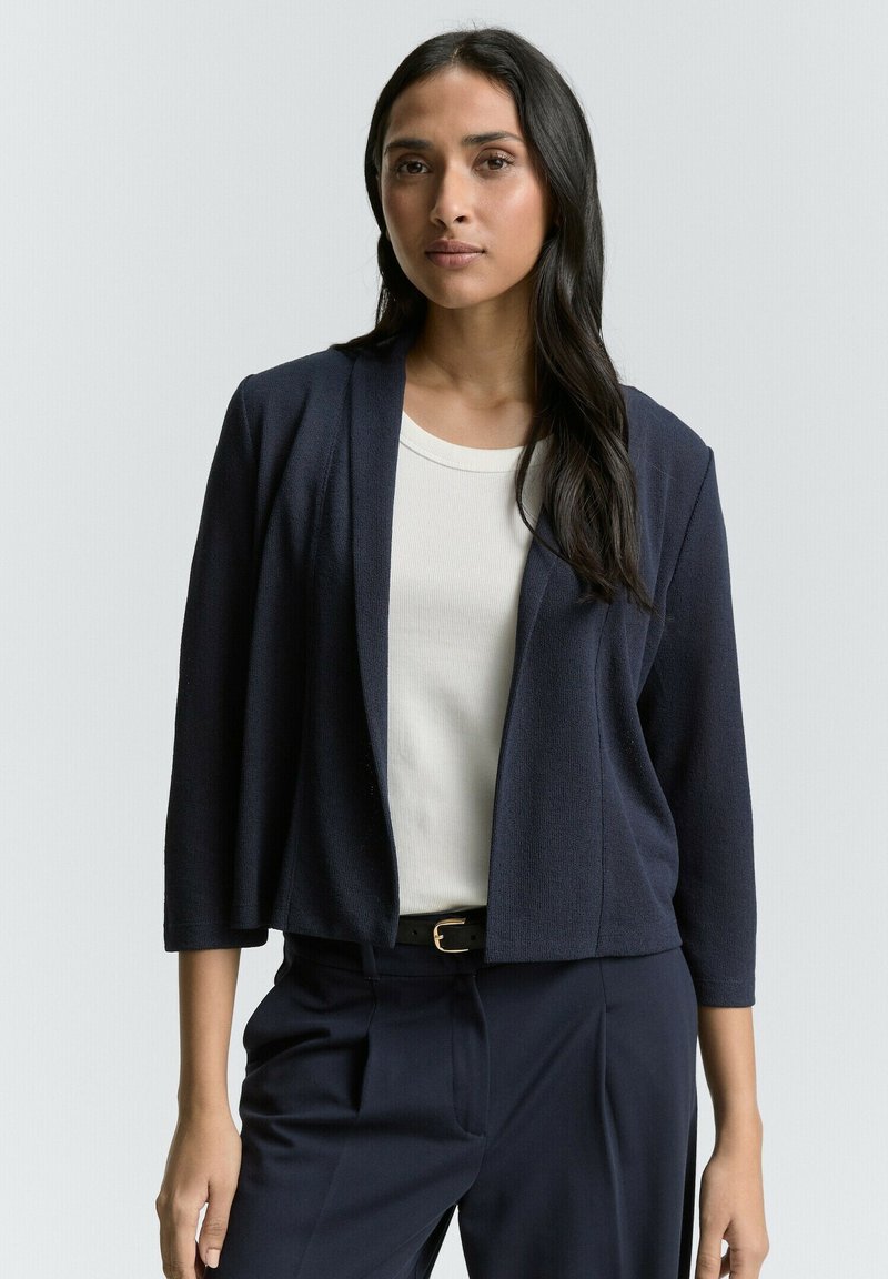 Blazer cropped di colore blu navy con maniche a tre quarti, tessuto texturizzato, design aperto davanti, abbinato a una maglietta bianca e pantaloni blu navy.