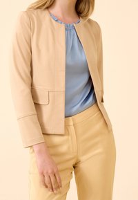 Veste beige ajustée avec un col rond et deux poches avant, associée à un chemisier bleu clair et un pantalon jaune. Texture du tissu lisse.