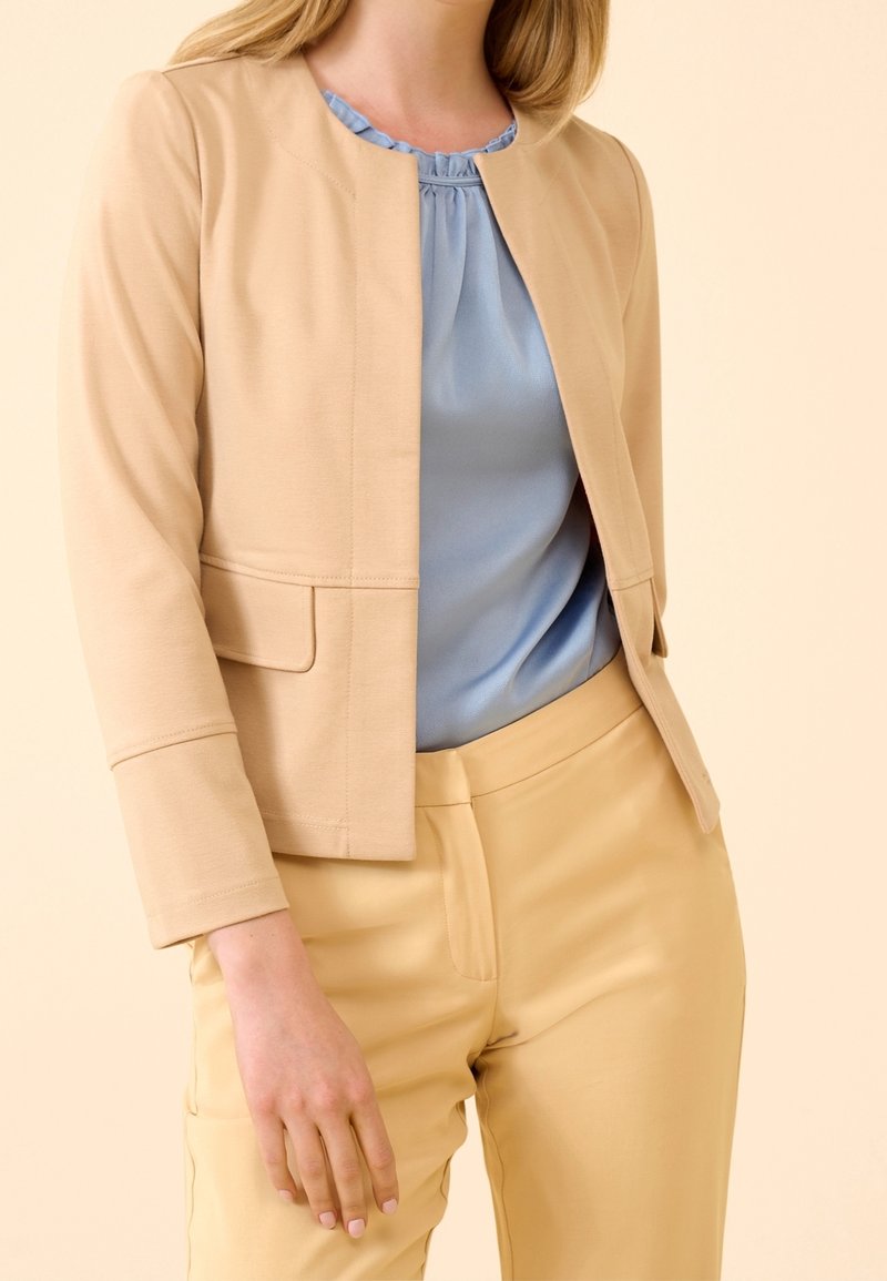 Veste beige ajustée avec un col rond et deux poches avant, associée à un chemisier bleu clair et un pantalon jaune. Texture du tissu lisse.