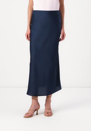 Jupe longue bleu marine en tissu lisse et léger avec une silhouette élancée. Associée à des sandales à talons hauts beiges à lanières.