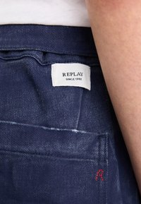 Donkerblauwe denim jeans met een zichtbaar wit merketiket met de tekst "REPLAY SINCE 1981." Beschikt over een rode geborduurde accent en een subtiele textuur.
