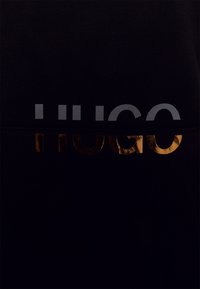 Sort stof med ordet "HUGO" i metallisk bronze og grå tekst, med en glat tekstur og minimalistisk design.
