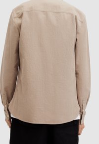 Camisa de manga larga beige con una tela texturizada, dobladillo curvado y puños abotonados, con un diseño sencillo y limpio sin patrones visibles.